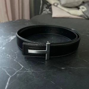 T0m F0rd bälte - Snyggt svart bälte med ett stilrent silverfärgat spänne. Perfekt för att ge en elegant touch till din outfit. Bältet har en klassisk design med vita sömmar längs kanterna.      AA. Size 100. Nyskick