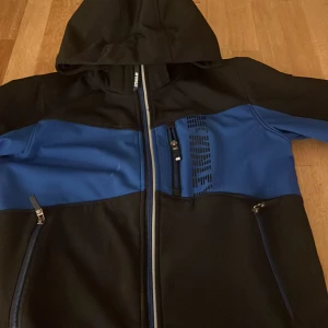 Svart och blå jacka från McKinley - Säljer en svart och blå jacka från McKinley med huva och dragkedja. Jackan har flera praktiska fickor med dragkedjor och en stilren design. Perfekt för utomhusaktiviteter och äventyr. 🏞️