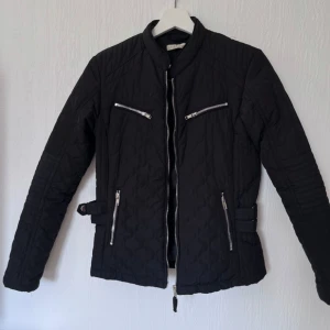 Svart jacka med dragkedjor - Snygg svart jacka med dragkedjor och bältesdetaljer i midjan. Jackan har en modern look med flera fickor och en stilren design.