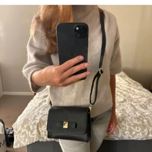 Svart axelväska med gulddetalj - Snygg svart axelväska med en elegant gulddetalj på framsidan. Väskan har en justerbar axelrem och en praktisk storlek för vardagsbruk. Perfekt för att bära dina nödvändigheter med stil.