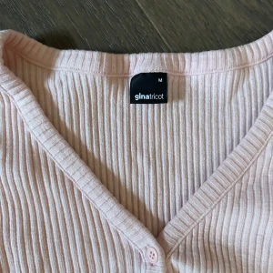 Ljusrosa ribbad kofta från Gina Tricot - Säljer en söt ljusrosa kofta från Gina Tricot i storlek M. Den är ribbad med en v-ringad design och knappar framtill. Perfekt för att antingen ha som en kofta eller bara en topp med knapparna stängda, då den är lite croppad! 