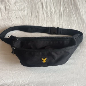 Svart Lyle & Scott midjeväska  - Snygg svart midjeväska med en gul logotyp framtill. Väskan har en justerbar rem och dragkedja för säker förvaring. Perfekt för att hålla dina saker nära till hands.
