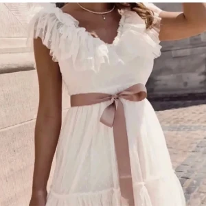 Studentklänning, Vit klänning med volanger  - Charmig vit klänning med volanger och ett fint prickigt mönster. Den har en v-ringad design och ett knytband i midjan (avtagbart) Perfekt för studenten