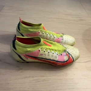 Färgglada fotbollsskor från Nike - Säljer ett par färgglada fotbollsskor från Nike med en iögonfallande design i gult, rött och svart. Skorna har en strumpliknande passform och är utrustade med dobbar för optimalt grepp på planen. Perfekta för den som vill sticka ut på fotbollsplanen!