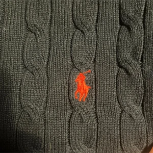 Mörkblå stickad tröja från Polo Ralph Lauren - Säljer en snygg mörkblå stickad tröja från Polo Ralph Lauren med det klassiska röda logotypbroderiet på bröstet. Tröjan har ett kabelstickat mönster och långa ärmar med ribbade muddar. Perfekt för kyliga dagar!