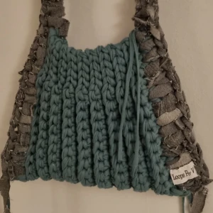 Virkad axelväska från Loops By V - Unik virkad axelväska i en härlig grön nyans med grå detaljer. Väskan är virkad i återvunnen textil samt återvunna jeans. Mkt rymlig modell. Perfekt för att ge din outfit en personlig touch. Mått: L ca 33cm H ca 22cm Axelrem ca 95cm.