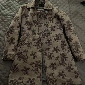 Blommig kappa från Vero Moda - Säljer en elegant kappa från Vero Moda i beige med brunt blommönster. Den har en dubbelknäppt design med stora knappar och långa ärmar. Perfekt för att ge en stilfull touch till din garderob.