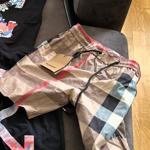Burberry rutiga shorts med snörning - Snygga shorts från Burberry med klassiskt rutigt mönster i beige, svart, rött och vitt. De har elastisk midja med svart snörning och är perfekta för sommaren. Lätta och bekväma, passar dig som vill sticka ut med stil.