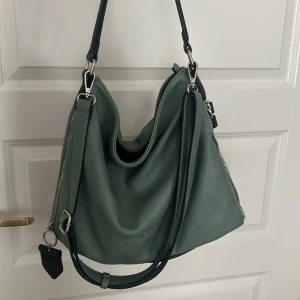 Grön axelväska från Picard  - Snygg grön axelväska från Picard med silverfärgade metalldetaljer och dragkedjor på sidorna. Väskan har både kort och lång rem, så du kan bära den på olika sätt. Perfekt storlek för allt du behöver ha med dig.