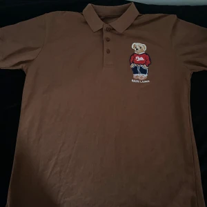 Brun pikétröja Polo Bear Ralph Lauren - Snygg brun pikétröja från Ralph Lauren med klassisk krage och knappar. På bröstet finns en broderad Polo Bear i röd tröja och blå byxor, samt Ralph Lauren-logga. Tröjan är i mjuk bomull och har korta ärmar. 