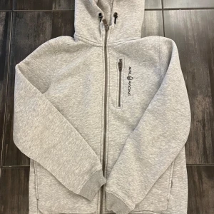 Grå hoodie från Sail Racing - Säljer en ljusgrå hoodie från Sail Racing med dragkedja framtill och huva. Tröjan har en bröstficka med dragkedja och logga på vänster sida. Mjuk insida och ribbade muddar vid ärmslut och nederkant.
