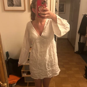 Vit linneklänning med v-ringning - Superfin vit kortklänning i luftig bomull och v-ringning både fram och bak. Klänningen har långa, vida ärmar och en avslappnad passform som ger en chill vibe. Perfekt för varma dagar eller när du vill känna dig extra fri. Endast använd 1 gång. Nypris ca 800kr