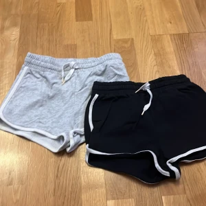 2-pack sportiga shorts med snörning - Två par sportiga shorts, ett par ljusgrå och ett par svarta, båda med vita kanter och snörning i midjan. De har elastisk midja och är tillverkade i mjuk bomullsmix, perfekta för träning eller chill. Klassisk retrostil med rundade benkanter och kontrastdetaljer.