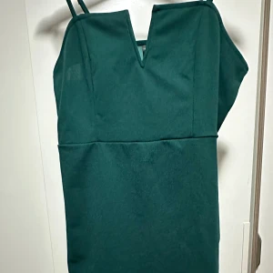 Mörkgrön kort klänning med v-ringning - Snygg mörkgrön klänning med smala axelband och markerad v-ringning. Modellen är kort och har en enkel, stilren design utan mönster. Materialet ser ut att vara polyester och klänningen har en liten slits i nederkanten för extra rörelsefrihet.