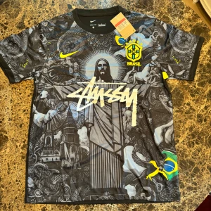 Nike x Stüssy Brasil fotbollströja L - Unik Brasilien fotbollströja från Nike och Stüssy med svart och grått konstnärligt mönster, stora tryck av Kristusstatyn och kyrkor, samt Brasil-logga och flagga. Kortärmad, Dri-FIT-material för bästa komfort. Stüssy-logga i vitt framtill och gula detaljer.