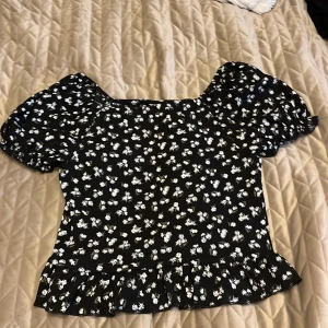 Svart blommig blus från Kappahl - Supersöt svart blus från Kappahl med vita blommor, puffiga korta ärmar och volangkant nertill. Blusen har en fyrkantig ringning och är gjord i ett lätt, skönt tyg som passar perfekt till jeans eller kjol. Perfekt för dig som gillar romantisk och trendig stil!