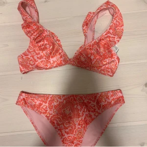 Rosa och röd bikini från Gina Tricot - Superfin bikini från Gina Tricot i rosa och rött med paisleymönster. Överdelen har breda axelband med volangdetaljer och byxan är klassisk i modellen. Perfekt för sommardagar vid stranden eller poolen! Aldrig använd! 