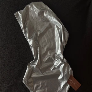 Silver bodysuit - Supersnygg bodysuit i metallic silver från SHEIN SKY. Modellen är högt skuren vid benen och har en glansig finish som verkligen sticker ut. Perfekt för dig som vill ha en trendig och unik look på stranden eller vid poolen.