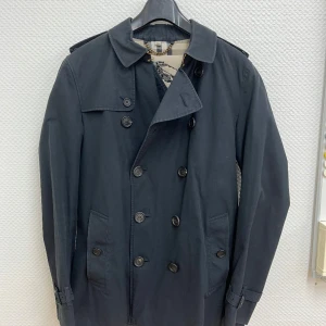 Burberry Men’s Heritage Kensington Short Trench Coat  - •	Storlek: 50 EU (motsvarar Medium–Large) 	•	Färg: Marinblå 	•	Modell: Kensington – kort längd 	•	Tillverkad i England 	•	Dubbelknäppning, axelklaffar, stormflik, justerbara ärmslut, bälte med metallspännen (D-ringar) och klassiskt rutigt Burberry-foder 	•	Skick: Mycket fint begagnat skick, sparsamt använd och väl omhändertagen 	•	Alla originaldetaljer finns kvar  Nypris ca 23 500 kr