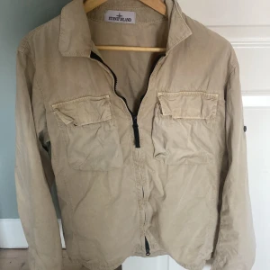 Beige overshirt från Stone Island - Snygg beige overshirt från Stone Island med två stora bröstfickor och dragkedja framtill. Jackan missar badgen men det går lätt att köpa för runt hundralappen. 