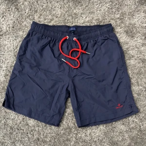 Marinblå badshorts från Gant - Snygga marinblå badshorts från Gant med röd snörning i midjan med en logga på ena benet. Shortsen har elastisk midja och en bakficka med kardborrestängning. Perfekta för stranden eller poolen. Byxorna har inga defekter och kostar 700 kr vid nypris. Använda fåtal gånger. 100% äkta badbyxor från Gant 