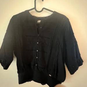 Svart blus med spets - Otrolig svart blus från Superdry med spetsdetaljer och knappar framtill. Blusen har trekvartsärm och spets längs ärmar och halsringning. Resår i midjan vilket ger en fin passform 