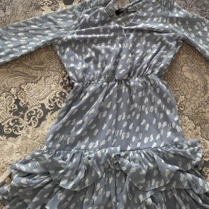 Blå mönstrad klänning Sisters Point - Supersöt blå klänning från Sisters Point med vitt abstrakt mönster, långa ärmar och volangdetaljer nertill. Klänningen har markerad midja och v-ringning, perfekt för dig som gillar en feminin och lekfull stil.