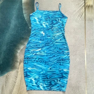 Blå kortklänning med vattenmönster - Snygg blå kortklänning med smala axelband och ett unikt vatteninspirerat mönster. Klänningen är figurnära och har en cool, modern vibe som passar perfekt för dig som vill sticka ut. Materialet känns mjukt och följsamt mot kroppen.