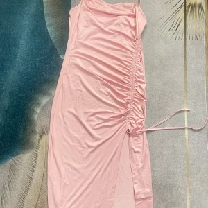Ljusrosa långklänning med snörning - Supersöt ljusrosa långklänning med smala axelband och snygg snörning på sidan som ger en rynkad effekt och slits. Perfekt för dig som vill ha en feminin och trendig look. Materialet känns mjukt och följsamt mot kroppen.