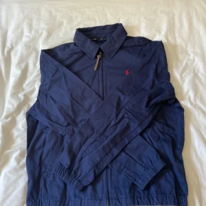 Blå vindjacka från Polo Ralph Lauren - Snygg marinblå vindjacka från Polo Ralph Lauren med klassisk krage, dragkedja och elastiska muddar. Jackan har röd broderad logga på bröstet och två fickor framtill. Perfekt för dig som vill ha en stilren och enkel jacka med ikonisk touch.