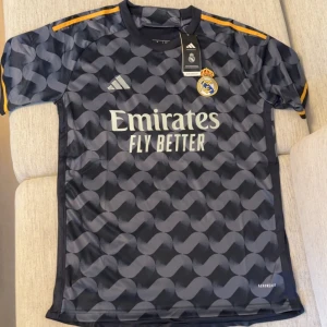 Real Madrid Adidas svart bortatröja - Snygg Real Madrid bortatröja från Adidas i svart med grått vågmönster och gula detaljer på axlarna. Tröjan har korta ärmar, klubbmärke på bröstet och 'Emirates Fly Better' tryckt framtill. Tillverkad i lätt och ventilerande material (Aeroready).