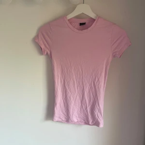 Ljusrosa t-shirt från Gina Tricot - En basic ljusrosa t-shirt från Gina Tricot i mjuk bomull. Modellen är figurnära med rund halsringning och korta ärmar. Perfekt att matcha med jeans eller kjol för en enkel och fräsch look.