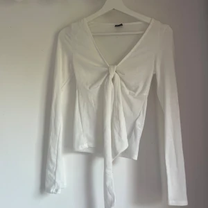 Vit långärmad knytblus Gina Tricot - Supersnygg vit långärmad topp från Gina Tricot i mjuk bomull. Djup v-ringning och knyt framtill ger en trendig look. Perfekt att styla med jeans eller kjol för en clean vibe.