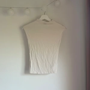 Vit polotopp från Gina Tricot XS - En stilren vit polotopp från Gina Tricot i storlek XS. Toppen har en hög hals och är ärmlös med en tight passform. Perfekt att styla med jeans eller kjol för en clean look. Gjord i mjukt bomullsmaterial.