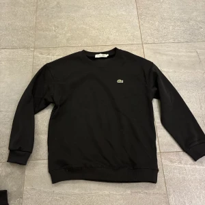 Svart sweatshirt från Lacoste - En stilren svart sweatshirt från Lacoste med klassisk rund hals och det ikoniska krokodilmärket broderat på bröstet. Tröjan har långa ärmar och ribbade muddar vid ärmslut och nederkant. Perfekt för en clean och avslappnad look.