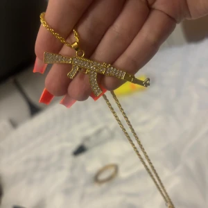 Guld halsband med AK47 -  halsband med ett AK47-hänge i guldfärg, täckt av glittrande stenar för extra bling. Kedjan är också guldfärgad och har en cool, urban vibe. Perfekt för dig som vill sticka ut med ett statement-smycke.