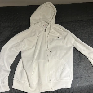 Vit hoodie från Lacoste med dragkedja - Snygg vit hoodie från Lacoste med dragkedja framtill och klassisk krokodillogga på bröstet. Tröjan har huva med snörning och två fickor framtill. Perfekt för en clean och sportig look.