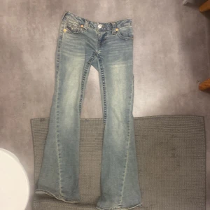 True Religion bootcut jeans ljusblå - Säljer ett par ikoniska ljusblå bootcut jeans från True Religion med snygga slitningar och klassiska kontrastsömmar. Låg midja och detaljerade bakfickor med knappar och logopatch. Perfekta för dig som vill ha en vintage vibe och cool passform.