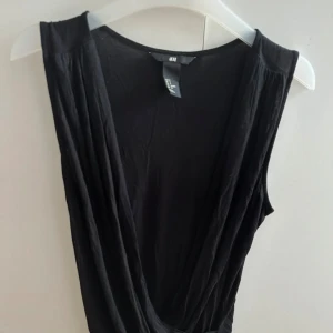 Svart omlott ärmlös topp H&M - Svart ärmlös topp från H&M med djup omlottringning framtill. Tunt och mjukt material som faller snyggt. Perfekt att styla med jeans eller kjol för en cool look
