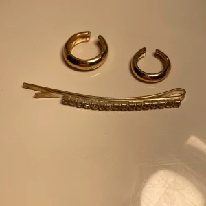 Guldiga örhängen och glittrigt hårspänne - Två chunky guldiga örhängen i creolstil och ett hårspänne med glittrande stenar. Örhängena har en enkel, rund form och hårspännet är dekorerat med små, klara stenar för extra bling. Perfekt för att lyfta din look med lite extra shine.