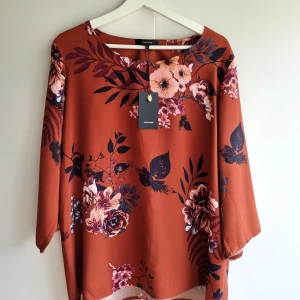 Blommig rostfärgad blus från Soyaconcept - Snygg blus i rostbrun nyans från Soyaconcept med stora blommiga mönster i lila, rosa och svart. Modellen har trekvartsärm och en lös passform. Perfekt för dig som gillar färg och mönster! Materialet känns mjukt och lätt.