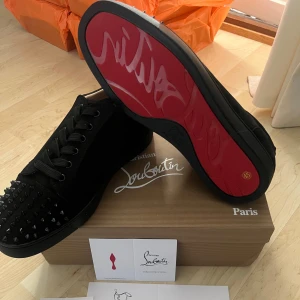 Christian Louboutin svarta sneakers med nitar - Svarta sneakers från Christian Louboutin i mocka med ikonisk röd sula och svarta nitar på tån. Skorna har snörning och rund tå, samt exklusiv känsla med guldfärgad logga på innersulan. Perfekta för dig som vill sticka ut med lyxig streetstyle.