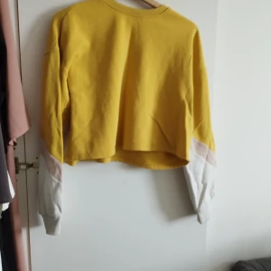 Gul croppad sweatshirt med vita ärmar - Snygg croppad sweatshirt i starkt gul färg med vita och ljusrosa detaljer på ärmarna. Tröjan har en loose passform och långa ärmar med muddar. Perfekt för dig som gillar färg och vill sticka ut lite extra.Kan hämtas upp i Malmö 