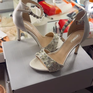 Catwalk vita klackpumps med pärlor - Eleganta vita pumps med öppen tå och smal klack från Catwalk. Skorna har en glittrig rem runt vristen och bred rem över tårna dekorerad med pärlor och blommiga detaljer. Perfekta för dig som vill sticka ut med en lyxig och feminin look.