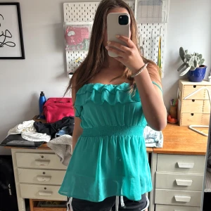 Turkos volangtop med smock i midjan - Supersöt turkos linnetopp med dubbla volanger upptill och smockad midja som ger en snygg siluett. Tunna axelband och lätt, flowigt tyg som känns luftigt. Perfekt för varma dagar och ger en fin färgklick till outfiten. Köpt secondhand men själv bara använt den en gång. Från H&M ursprungligen. Storlek 40 men passar mindre har själv storlek 36. 