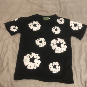 Svart t-shirt från Denim Tears S - Svart t-shirt från Denim Tears med unika vita blommönster tryckta över hela plagget. Klassisk passform med rund hals och korta ärmar. Perfekt statement-piece för dig som vill sticka ut med cool design och streetwear-vibe.