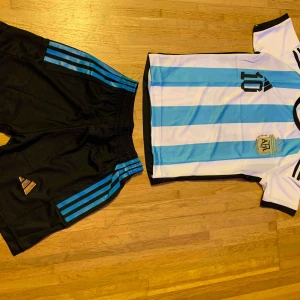 Argentina Messi fotbollströja set - Säljer ett Argentina fotbollströja och shorts set från Adidas. Tröjan har klassiska blå och vita ränder, nummer 10 och Messi-tryck på ryggen. Korta ärmar och broderad landslagsemblem. Shortsen är svarta med blå ränder och Adidas-logga. Perfekt för fotbollsälskare!