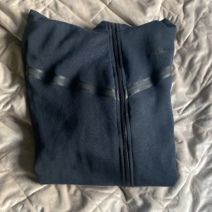 Svart Nike tech fleece hoodie - Snygg svart hoodiejacka från Nike med hel dragkedja framtill och diskreta svarta ränder över bröstet. Jackan har huva och är tillverkad i mjukt bomullsmaterial, perfekt för chill eller träning. Klassisk Nike-logga på bröstet och långärmad modell.