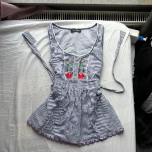 Randig blus med broderi från Perfectly Petite - Supersöt blus i blåvitt randigt från Perfectly Petite. Broderade blommor i färgglada toner framtill, knappar och knytband i midjan för extra passform. Ärmlös modell med lätt utställd nederdel och fina virkade detaljer vid halsringningen.