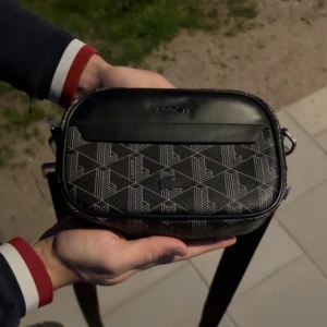 Svart axelväska från Lacoste - Säljer en svart axelväska från Lacoste med geometriskt mönster i vitt. Väskan har en stilren rektangulär form, justerbar axelrem och dragkedja upptill. Tillverkad i skinnliknande syntetmaterial med diskret Lacoste-logga framtill. Perfekt för att bära det viktigaste.